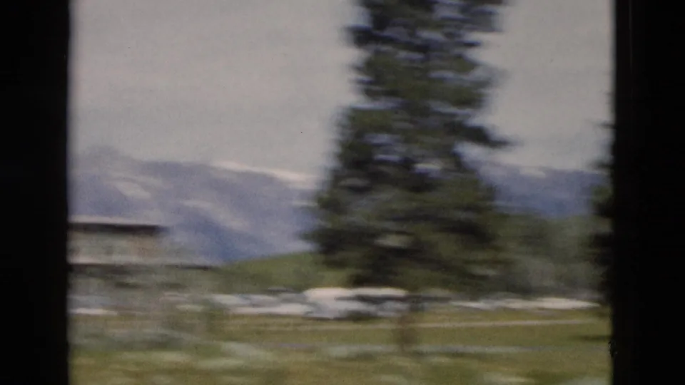 Handheld 8mm pan across Yellowstone lodge grounds, with a blurred evergreen, cars, grass, and mountain backdrop.