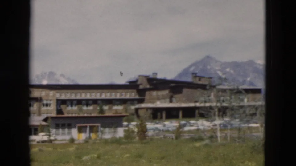 1958 Yellowstone frame showing a multi-story rustic lodge with grass, trees, parked cars, and mountains beyond.