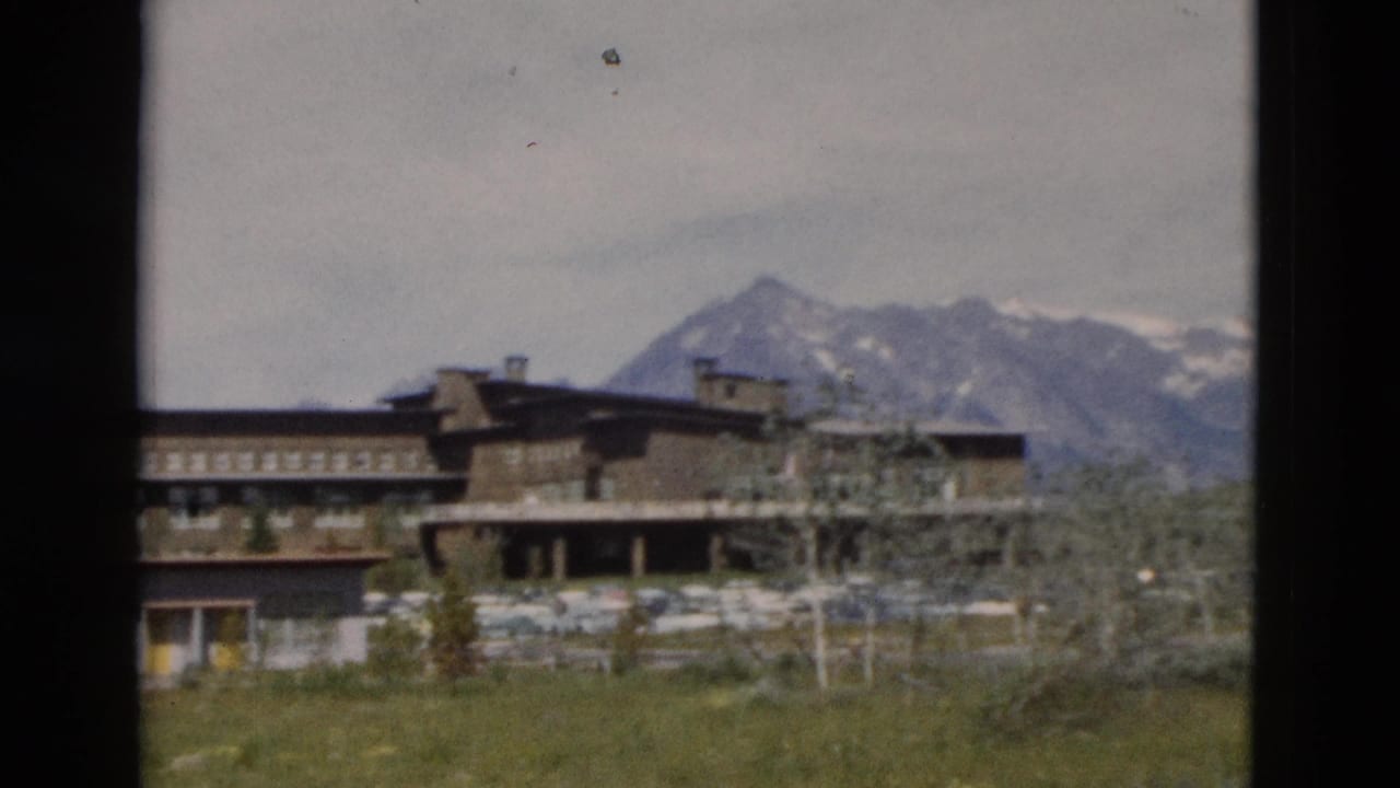 Lodge-style building in front of snow-capped mountains and open grass.