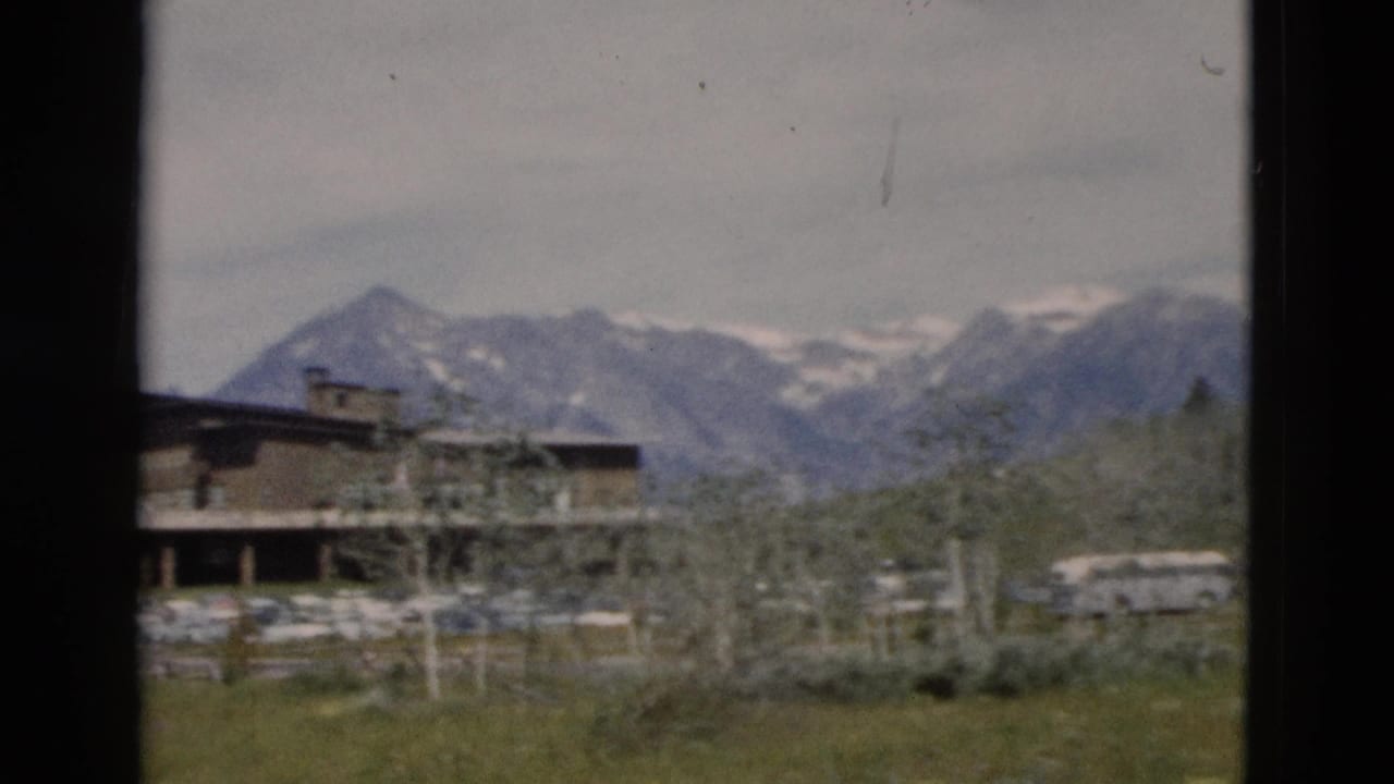 1958 Yellowstone-area view of a lodge exterior, parked vehicles, trees, and a distant snow-streaked mountain range.