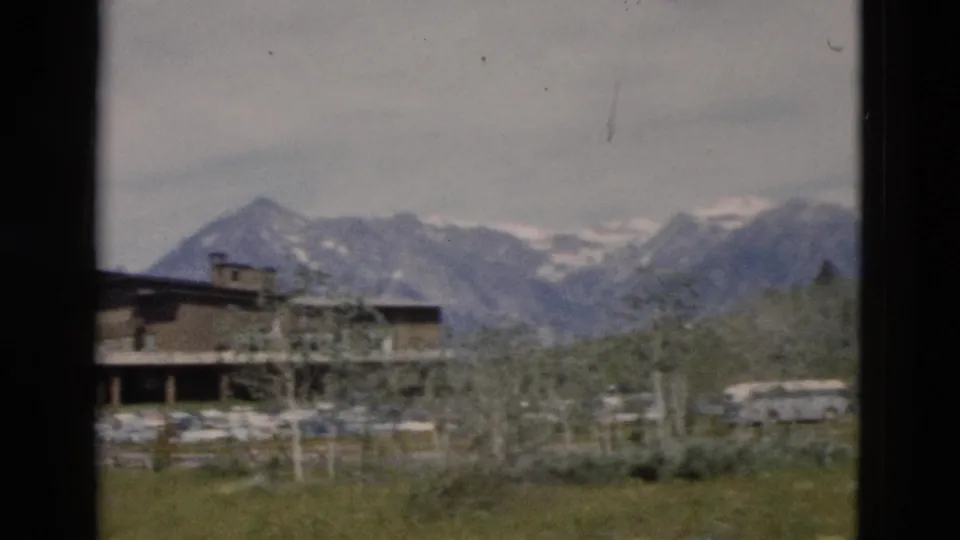 1958 Yellowstone-area view of a lodge exterior, parked vehicles, trees, and a distant snow-streaked mountain range.