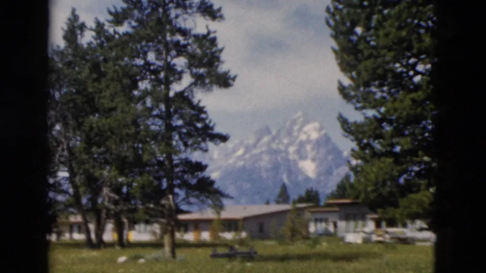 1958 Yellowstone-area frame showing evergreens, a low lodge or motel building, lawn, and a snow-capped mountain peak.