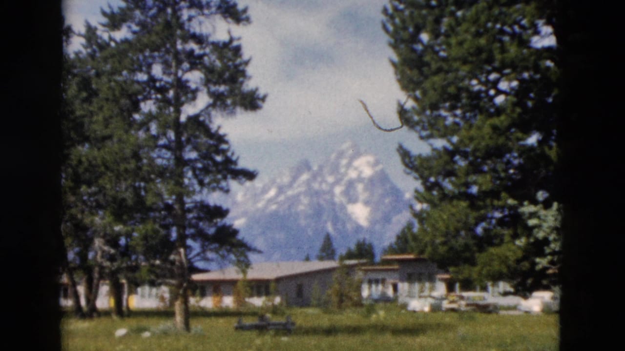 1958 Yellowstone-area home-movie frame showing snow-capped mountains, evergreens, a lawn, and low visitor buildings.