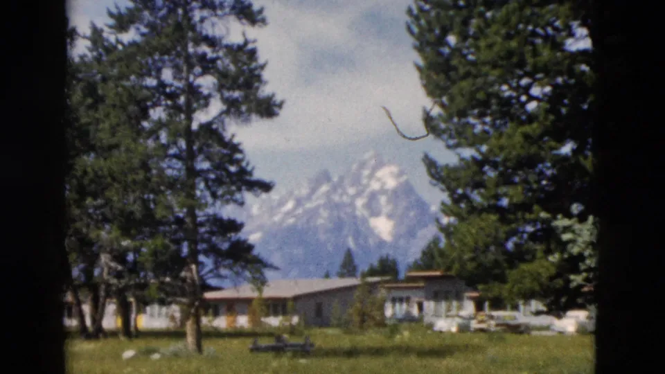 1958 Yellowstone-area home-movie frame showing snow-capped mountains, evergreens, a lawn, and low visitor buildings.