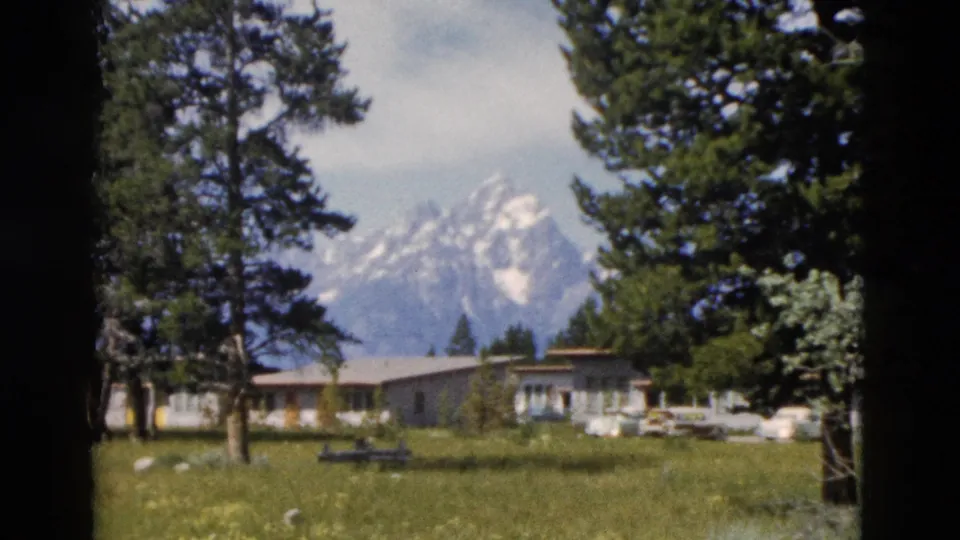 1958 Yellowstone-area frame showing a lawn with a picnic table, evergreens, low buildings, cars, and snow-capped peaks.