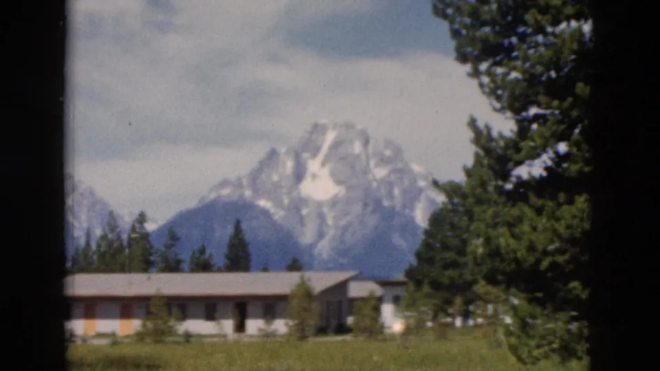 1958 Yellowstone-area home-movie view of snowy mountain peaks beyond a low lodge-like building and surrounding evergreens.