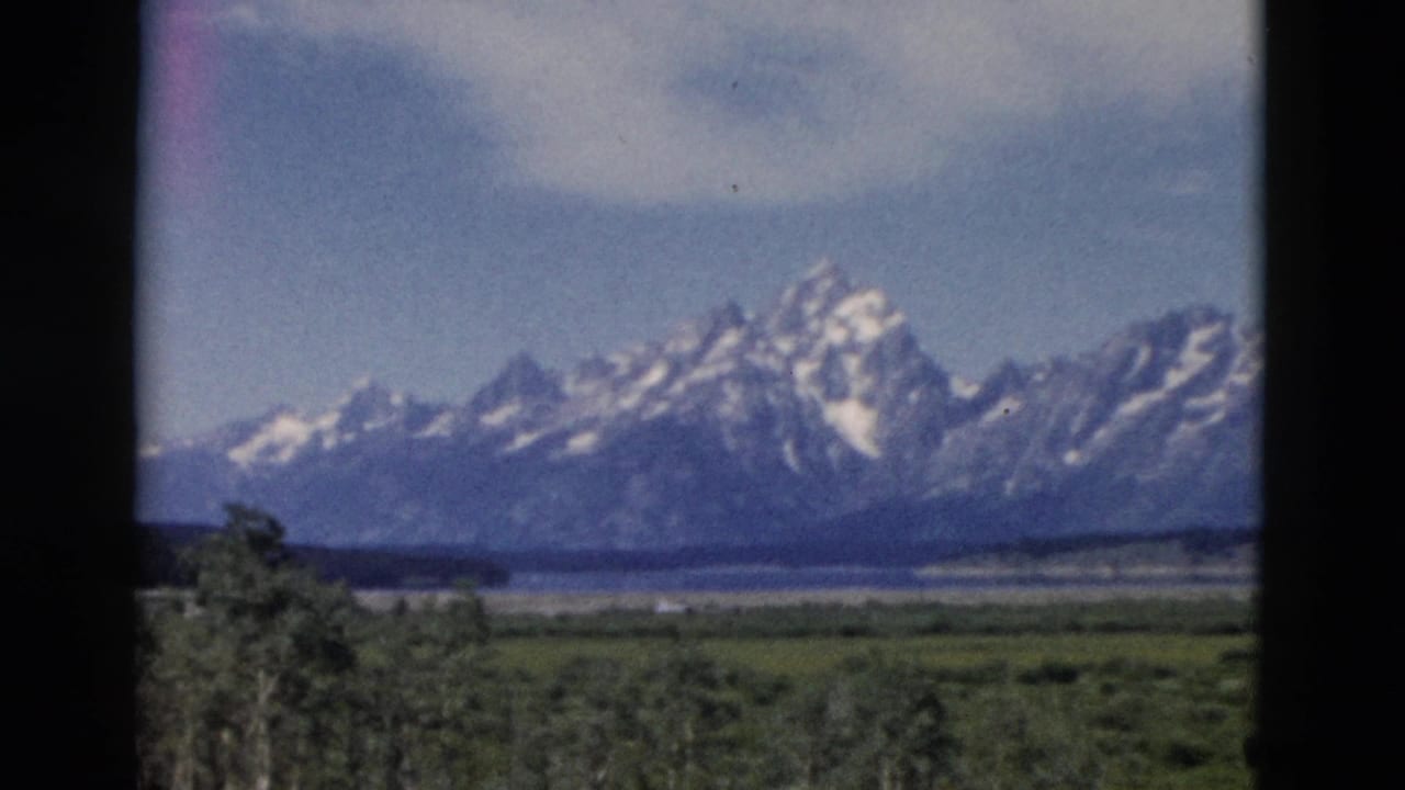 1958 Wyoming home-movie frame showing a snow-capped mountain range beyond meadow and water, bordered by dark window-like edges.
