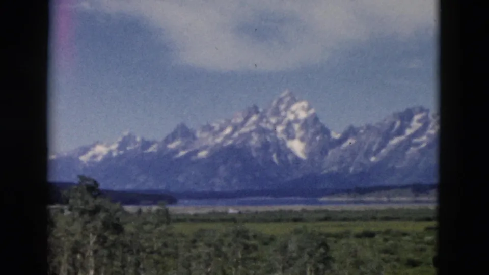 1958 Wyoming home-movie frame showing a snow-capped mountain range beyond meadow and water, bordered by dark window-like edges.