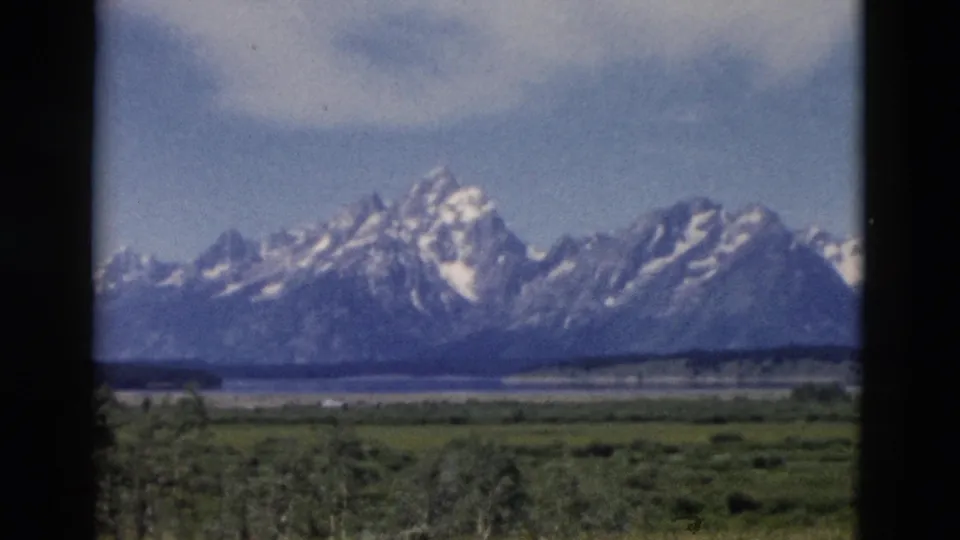 Soft 8mm frame of Wyoming mountain scenery, with snow-streaked peaks, meadow, waterline, trees, and sky visible.