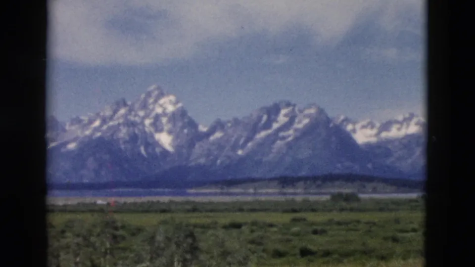 1958 8mm Yellowstone-area mountain vista with snow-capped peaks, meadow, waterline, trees, and dark window-like borders.
