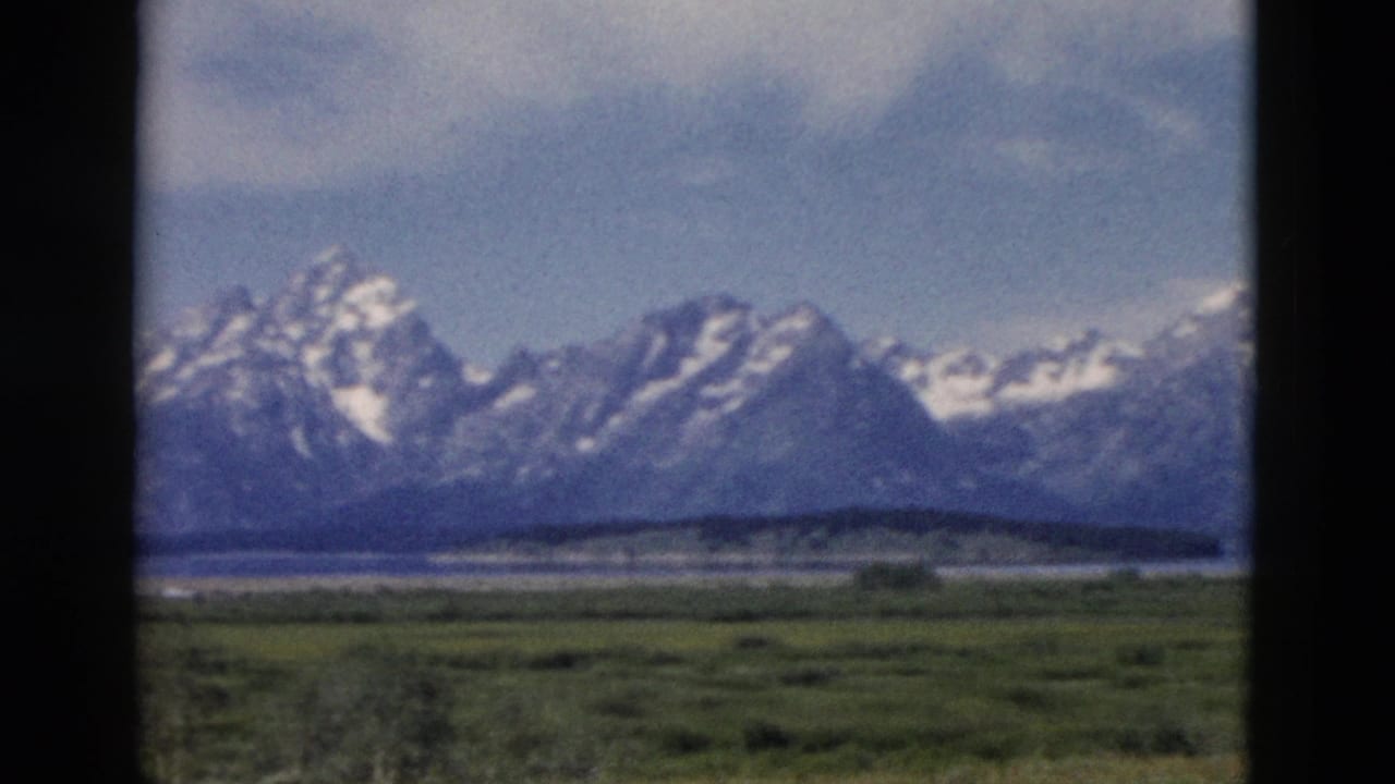 1958 8mm Yellowstone-area mountain view with snowy peaks, a green valley, distant water, and dark window-like borders.