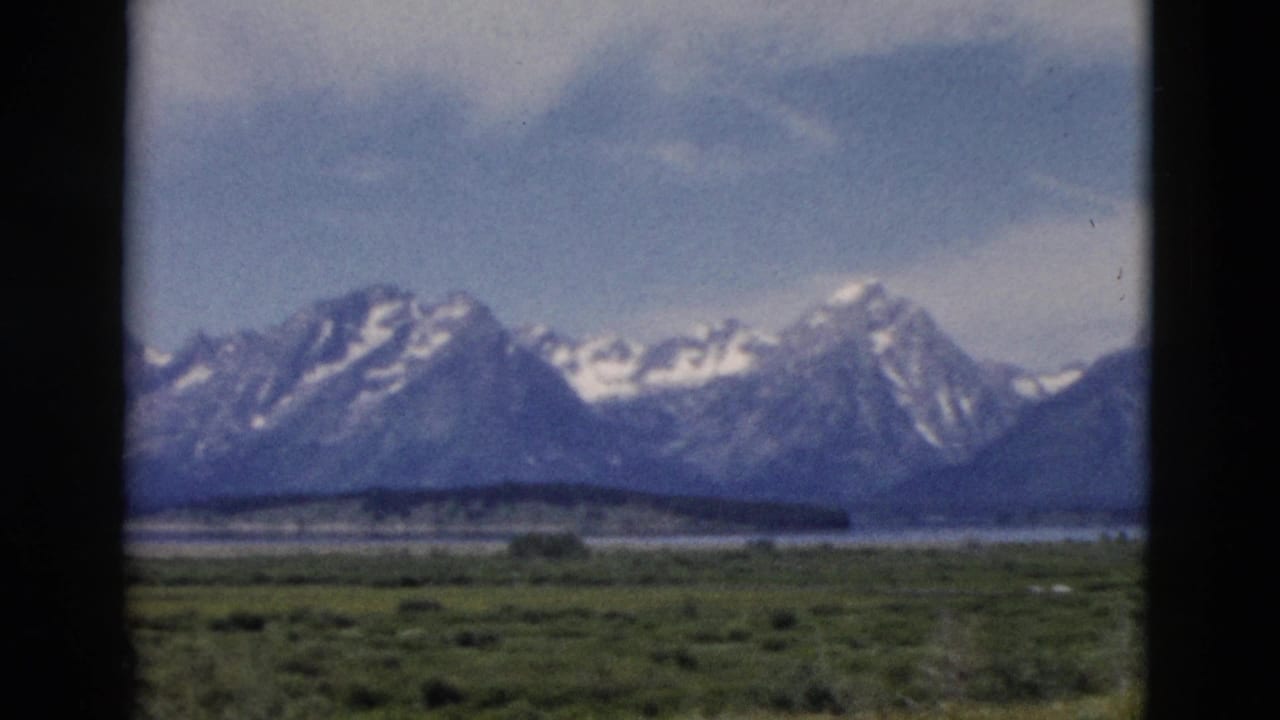 Mountain peaks with snow patches beyond low green terrain and a calm strip of water.