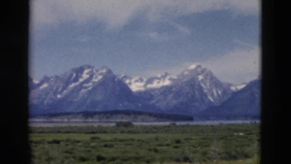 Vintage 8mm view of Wyoming mountain scenery, with snow-streaked peaks, green foreground, water, and dark framing edges.