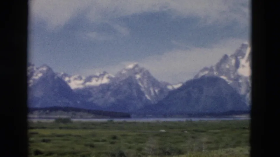1958 home-movie frame of Yellowstone-area peaks, grassland, distant water, and a darker rounded mountain in soft 8mm color.