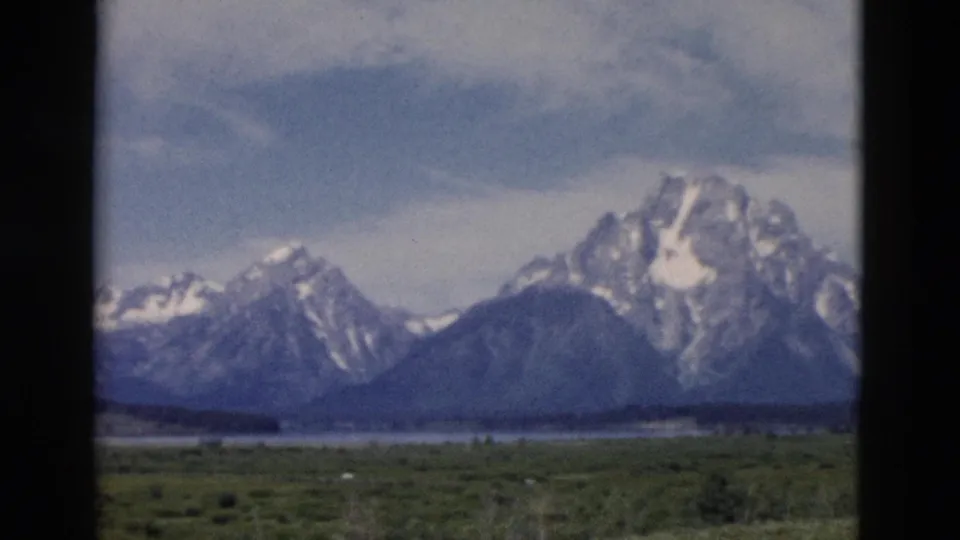 Soft 1958 8mm Yellowstone-area landscape with snowy peaks, dark slopes, green valley, and a muted cloudy sky.