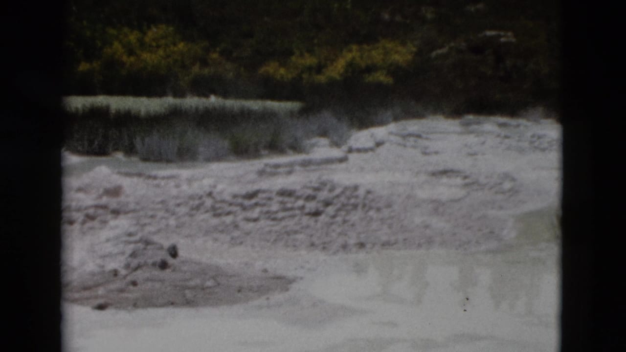 1958 Yellowstone 8mm view of a pale rocky geothermal surface, shallow mineral water, small stones, and dark vegetation beyond.