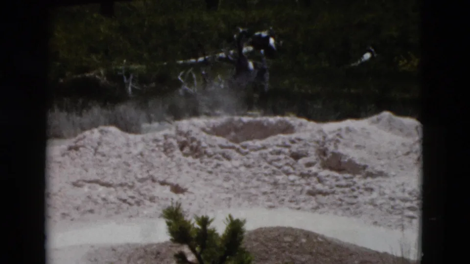 1958 Yellowstone 8mm frame showing steam rising behind a pale geothermal mound, with trees, shrubs, and shallow mineral water visible.