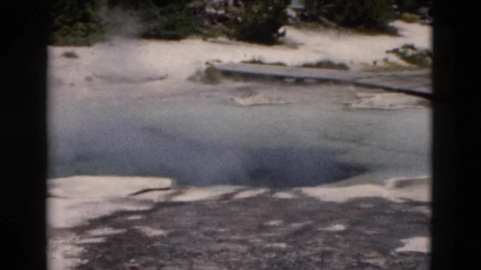 Wide Yellowstone home-movie frame showing a steaming, partly frozen pond bordered by snow patches and dark evergreens.