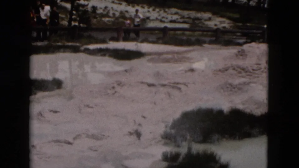 1958 Yellowstone home-movie frame showing visitors on a boardwalk beside pale mineral ground, shrubs, and evergreen trees.