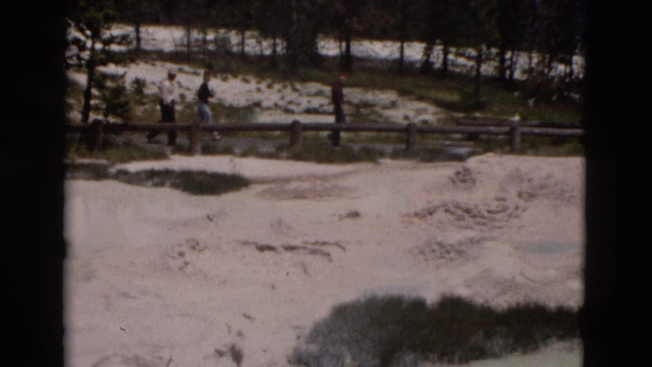 1958 Yellowstone frame with distant visitors crossing a boardwalk over a light-toned geothermal area bordered by trees.