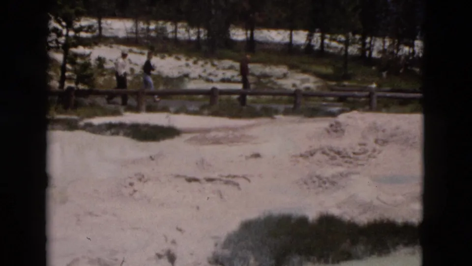 1958 Yellowstone frame with distant visitors crossing a boardwalk over a light-toned geothermal area bordered by trees.