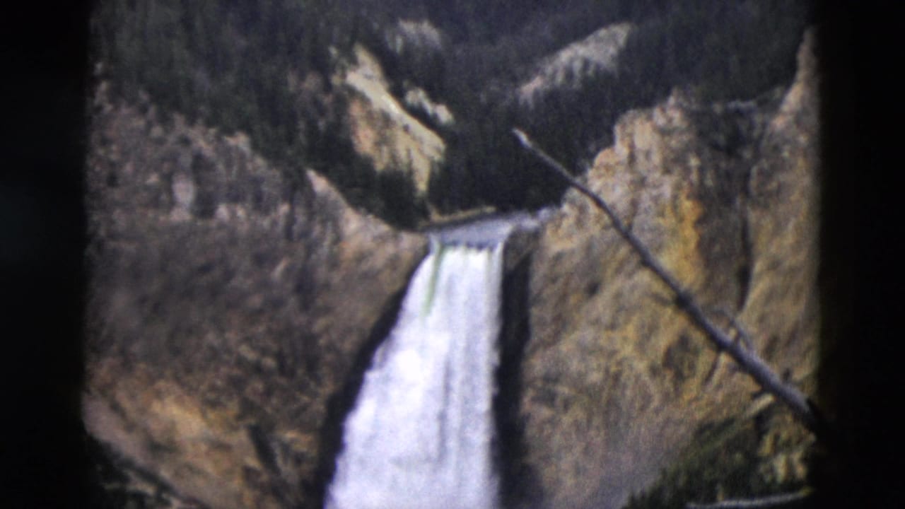 Waterfall dropping from a rocky canyon rim with dark forested slopes above.