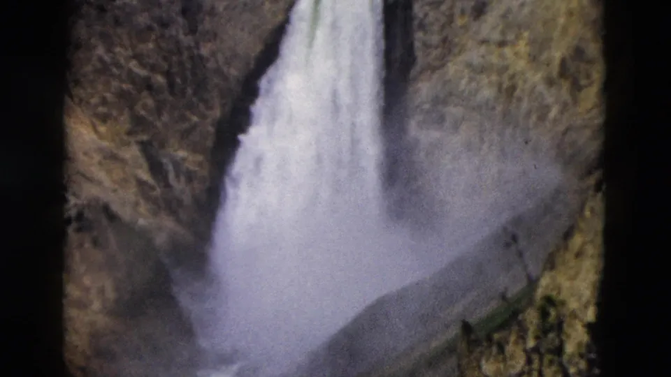 1958 8mm Yellowstone frame showing a tall waterfall descending into a mist-filled canyon bordered by rugged rock slopes.