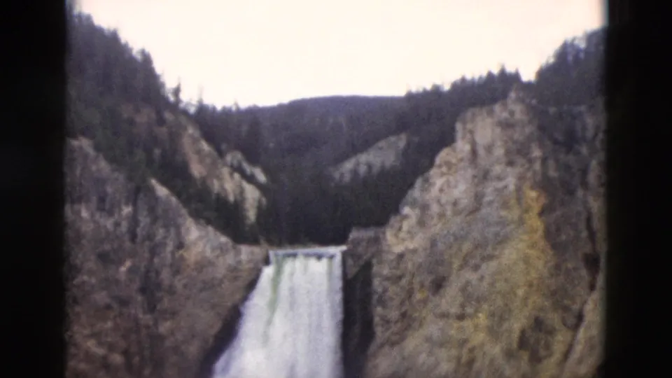1958 Yellowstone home-movie frame showing a waterfall between steep canyon walls and dark pine-covered slopes.
