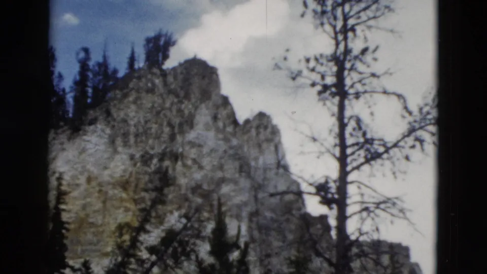 1958 Yellowstone-area mountain view with a pale rock peak, evergreen forest, bare branches, blue sky, and soft cloud on 8mm film.