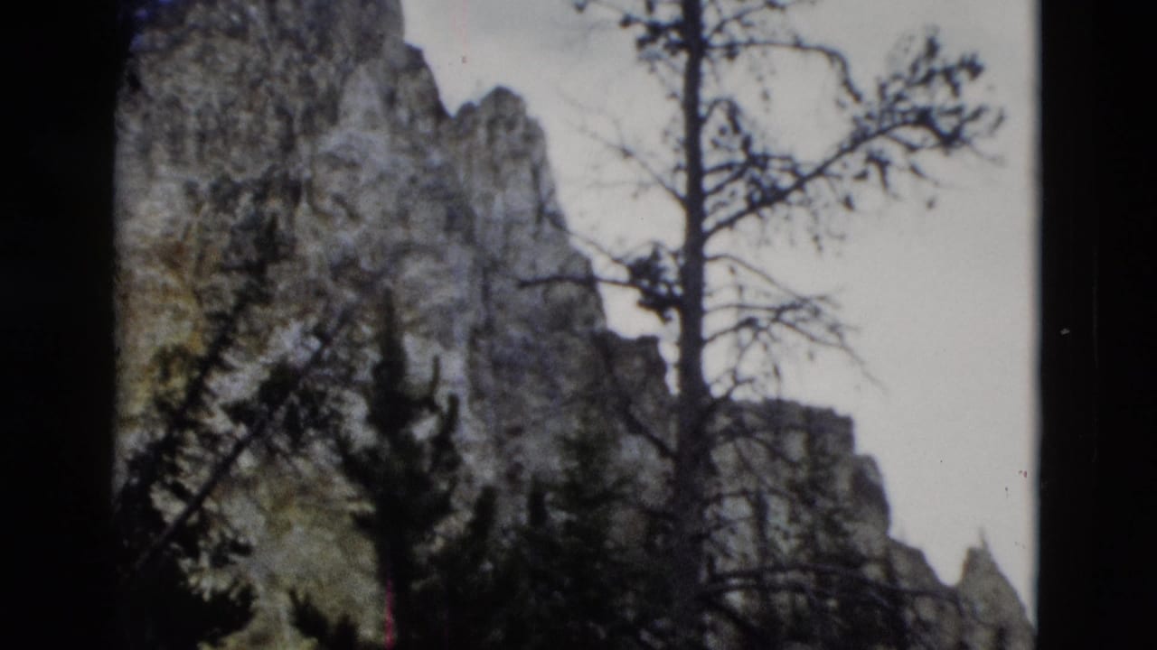 1958 Yellowstone 8mm frame showing gray rock formations, evergreen silhouettes, bare branches, and a pale sky.
