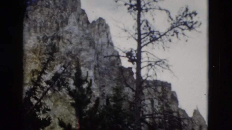 1958 Yellowstone 8mm frame showing gray rock formations, evergreen silhouettes, bare branches, and a pale sky.