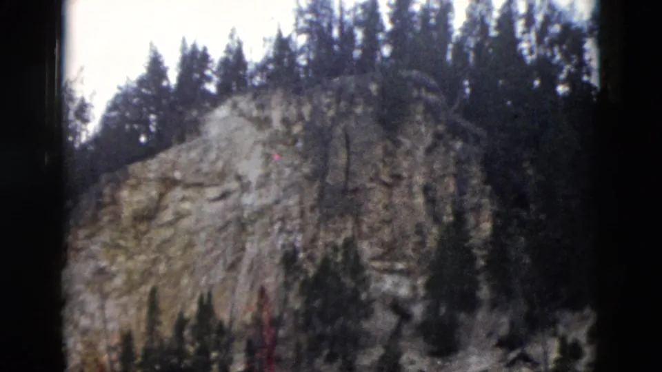 1958 Yellowstone 8mm view of a rugged cliff face with evergreen trees along the ridge and darker trees in the foreground.