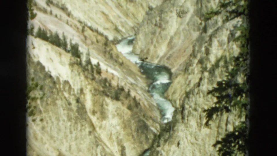 1958 Yellowstone home-movie frame showing a winding river at the bottom of a steep canyon with pale rock walls and scattered evergreens.
