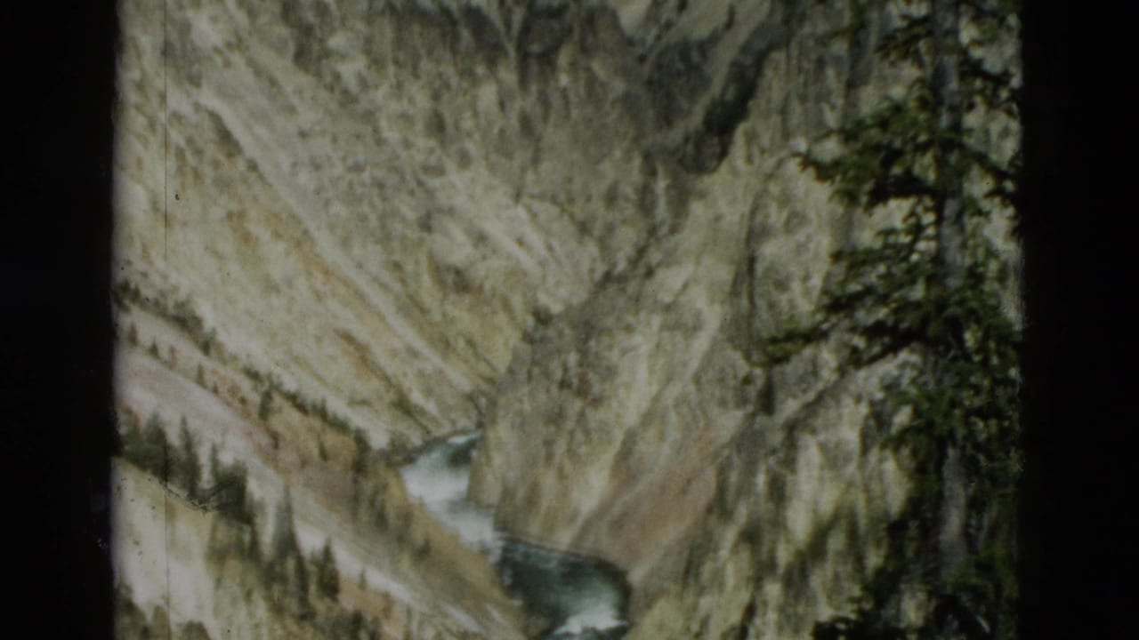 1958 Yellowstone home-movie frame of a narrow river channel below layered canyon walls, with dark evergreen branches along the right edge.