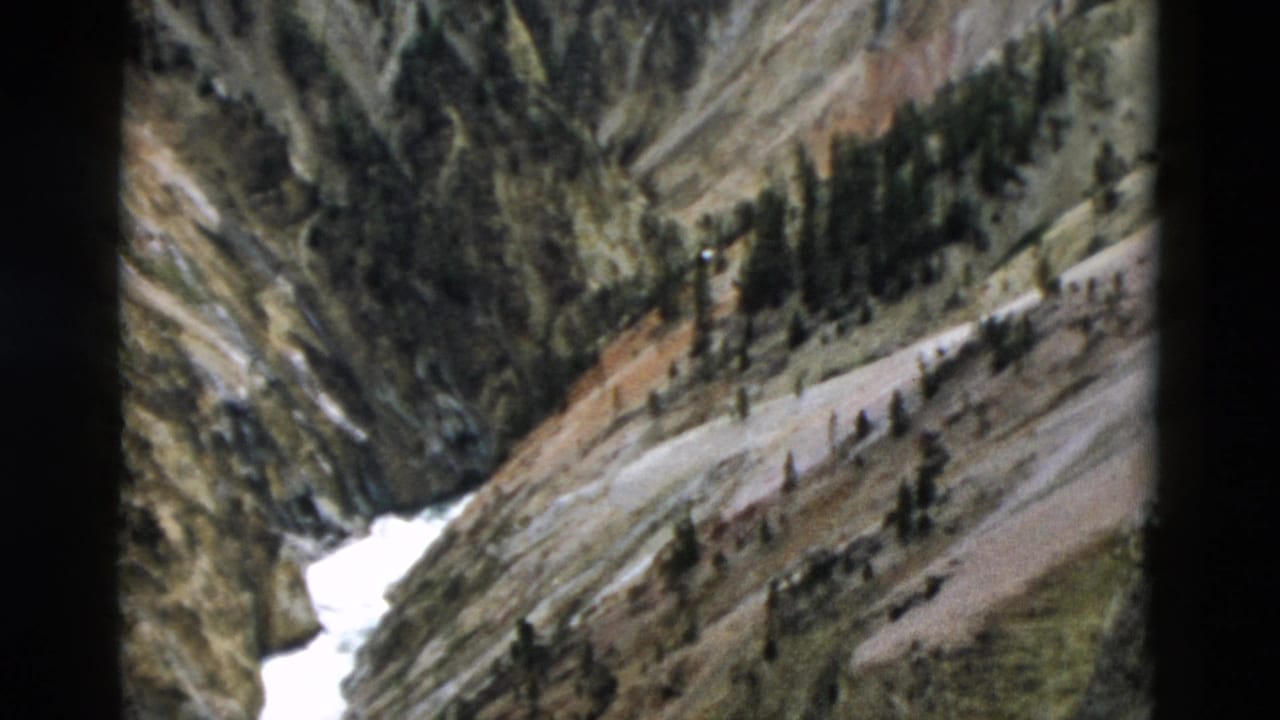 Yellowstone, Wyoming frame from 1958 with a distant snowmelt channel, rugged slope, conifers, and a pale roadlike line.