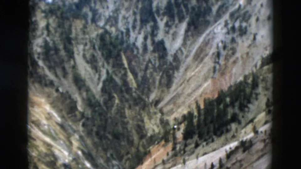 Soft 8mm Yellowstone frame from 1958 showing a canyon hillside, conifers, exposed rock, and a pale roadlike strip.