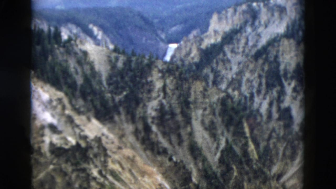 1958 Yellowstone home-movie frame showing a canyon, evergreen forest, rocky cliffs, and a distant waterfall.