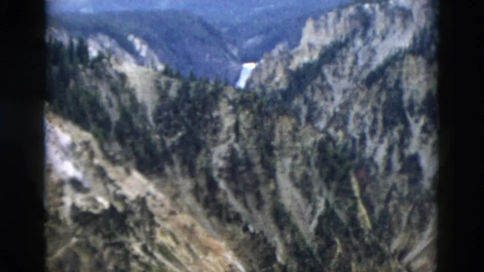 1958 Yellowstone home-movie frame showing a canyon, evergreen forest, rocky cliffs, and a distant waterfall.