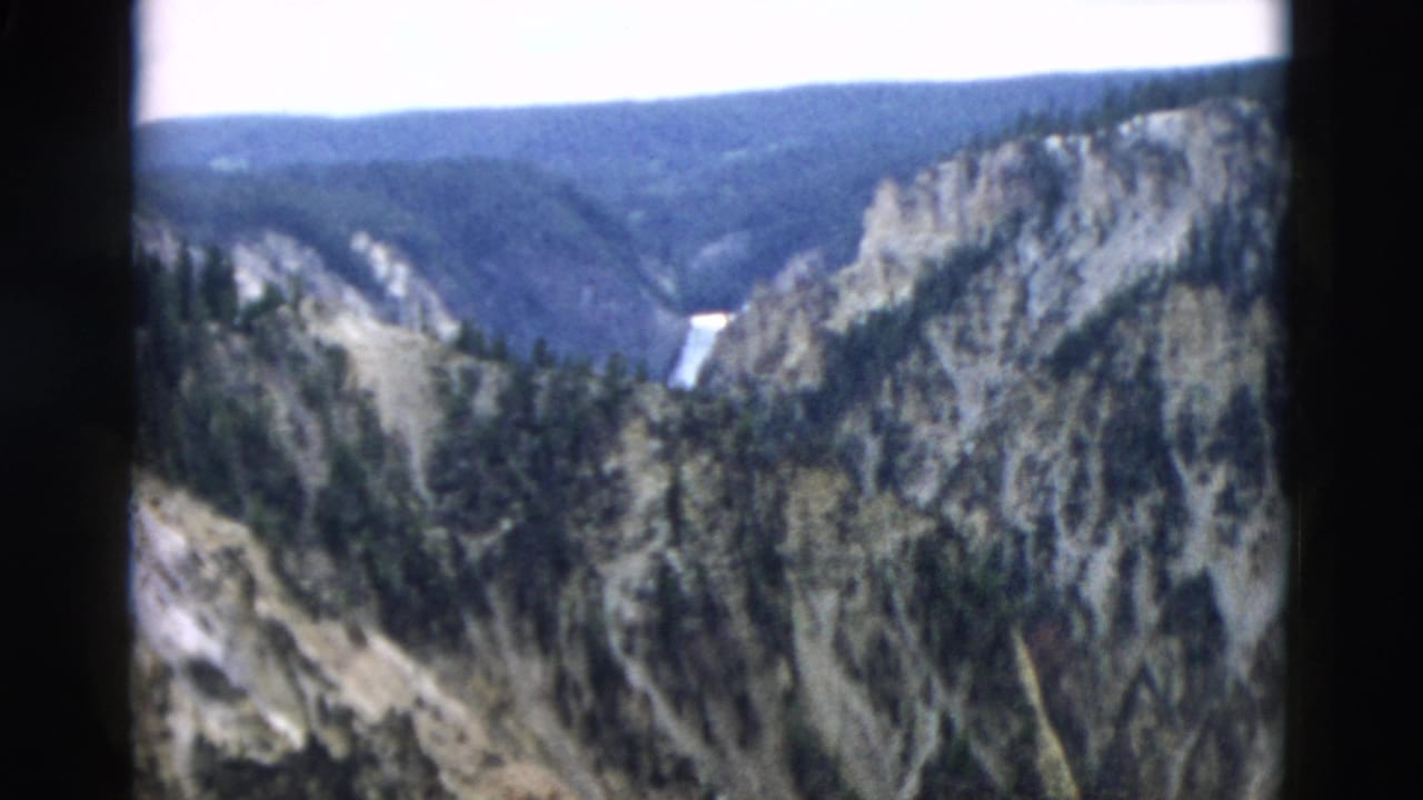 Wider canyon view with cliffs, dark evergreen trees, and a waterfall in the distance.