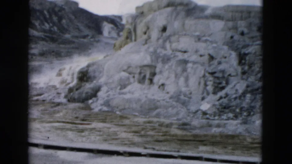 1958 Yellowstone 8mm frame showing pale geothermal terraces, mineral ledges, wet ground, and darker hillside forms beyond.