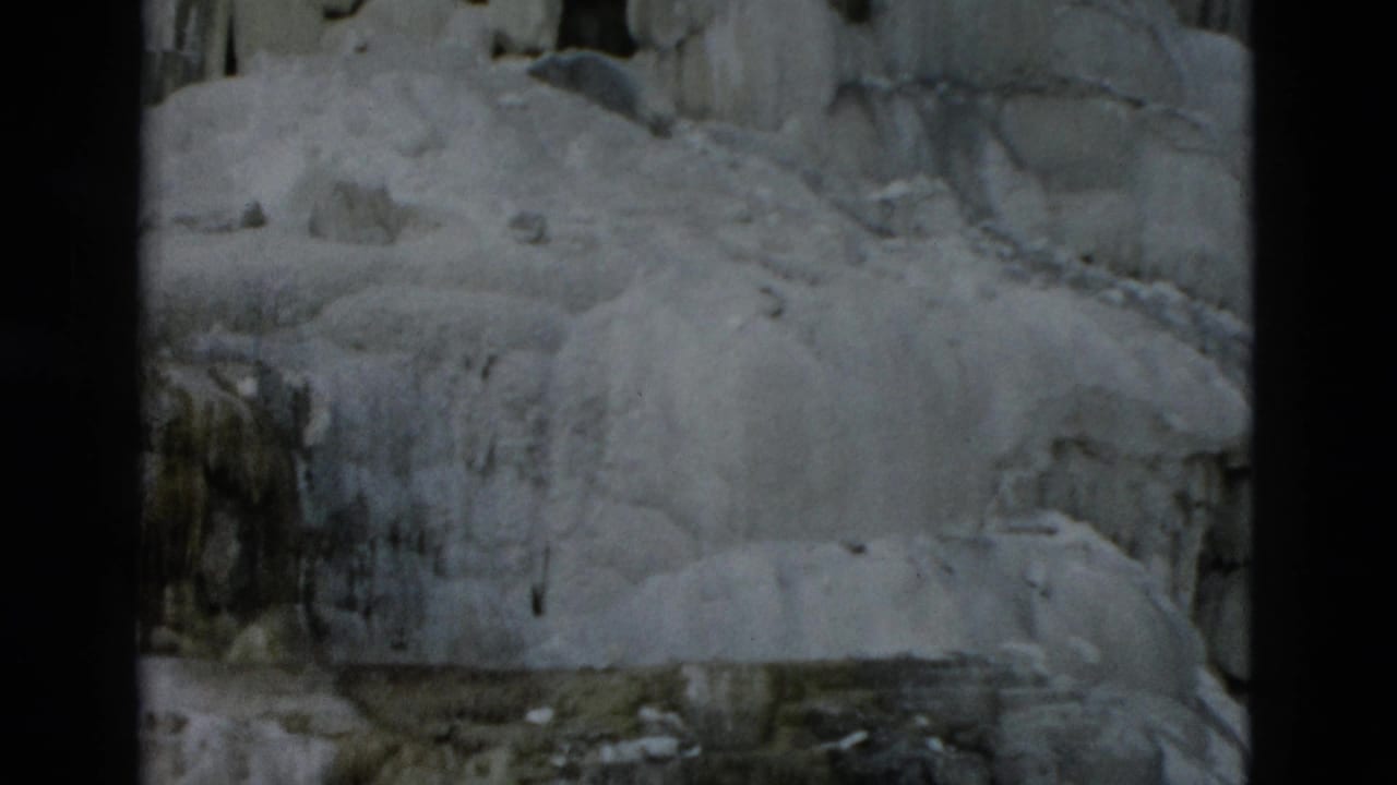 Frozen rock face with pale ice sheets, hanging icicles, and dark 8mm frame borders.