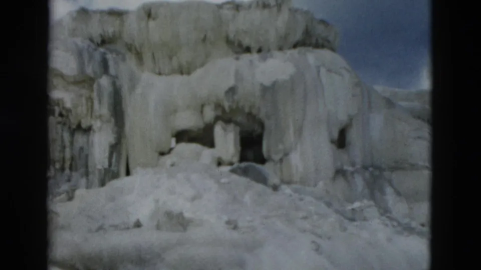 1958 Yellowstone frame showing icy rock, a shadowed opening, snow buildup, and blue-green water at the base.