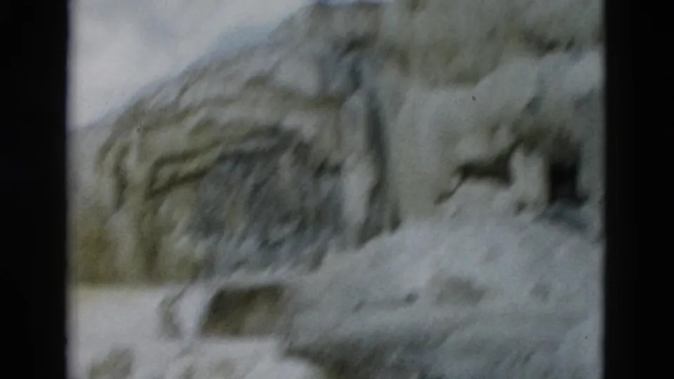 Soft-focused Yellowstone home-movie frame with pale ice over rock, dark crevices, and a partially visible pool below.