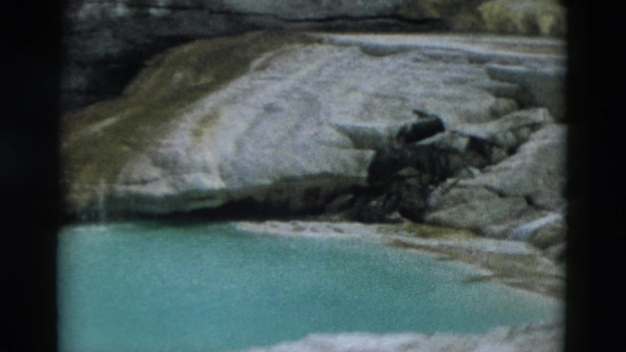 1958 Yellowstone 8mm frame showing a turquoise pool, pale terrace rock, and dark vegetation near the basin edge.