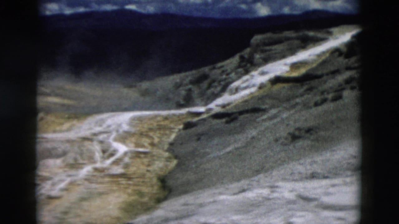 1958 Yellowstone 8mm frame showing pale geothermal terraces, steam, rocky slope, and dark clouded sky.