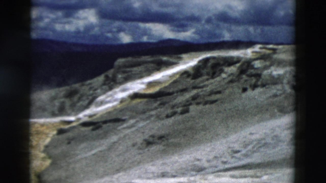 1958 Yellowstone view of a pale thermal stream crossing rocky terraces below a dark mountain ridge and clouded sky.