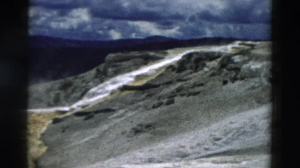 1958 Yellowstone view of a pale thermal stream crossing rocky terraces below a dark mountain ridge and clouded sky.