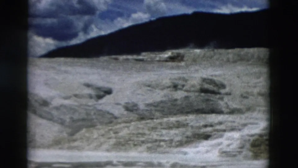 1958 Yellowstone 8mm frame showing pale mineral flats, scattered rocks, distant ridge, and heavy clouds.