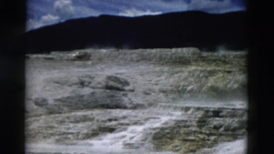 1958 Yellowstone frame showing mineral flats, a pale thermal runoff channel, distant ridge, and clouded sky in soft 8mm color.