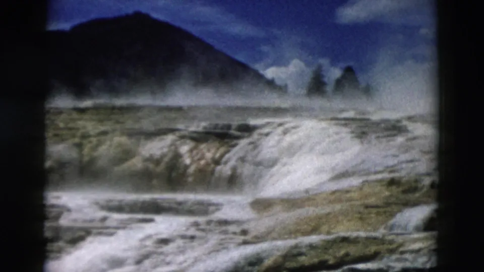 Visitor-shot 1958 Yellowstone view of a steaming mineral terrace edge with hills and clouded sky beyond.